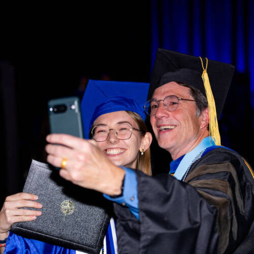 faculty member on right next to a new graduate taking a selfie during the December commencement at Van Andel Arena